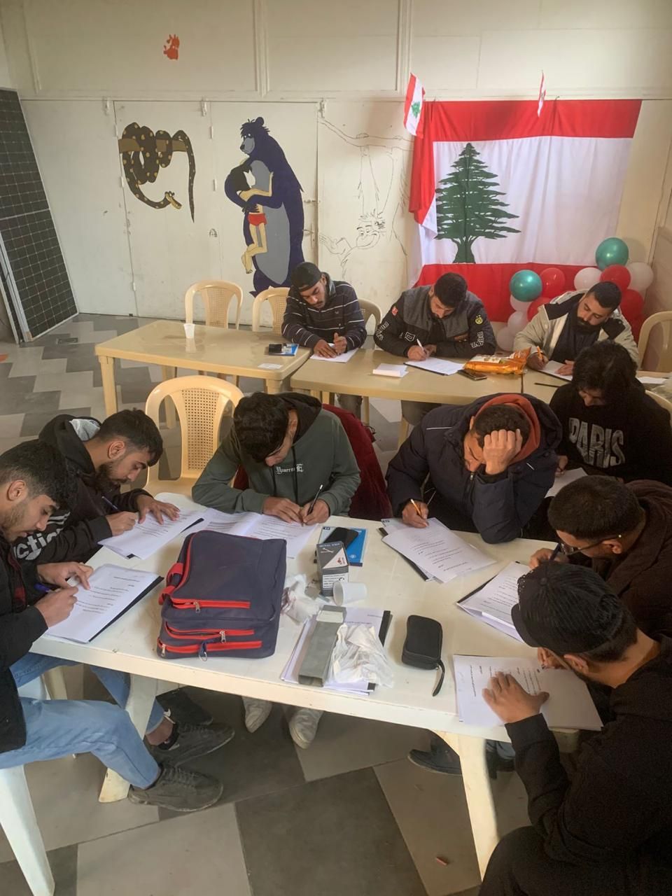 متدرّبون جالسون حول طاولة يدوّنون ملاحظاتهم خلال دورة الطاقة الشمسية، وخلفهم علم لبنان